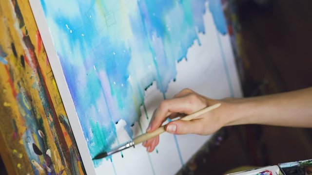 Young woman artist draw pictrure with watercolor paints and brush on easel canvas closeup hand
