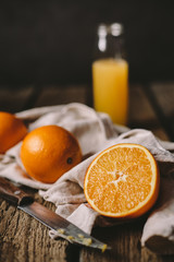 Obraz na płótnie Canvas Big fresh juicy orange and a half of one lie on the old crumpled kitchen towel, on the rough wooden table, old knife, glass bottle with tasty freshly-squeezed orange juice and the dark gray background