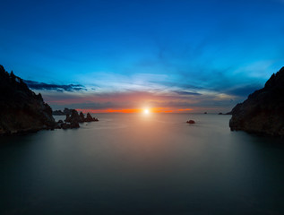 Long exposure seascape at sunset\sunrise. The sun goes down, surrounded by a halo and clouds. View of the cliff into the sea and distant islands. The backlight sunbeam. Paleokastrica. Corfu. Greece.