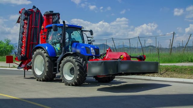 A blue tractor is standing still on a road. There is a trailor connected to it. It is a nice summer day in a village.
