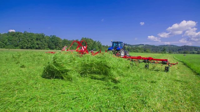 A farmer is driving a big tractor on the grass field. He is cutting grass with a grass cutting machine. Hay is flyng everywhere around. It is a nice summer day.
