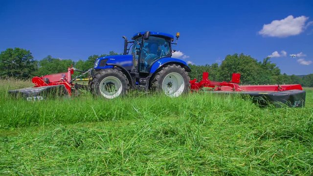 A big blue tractor is slowly driving on the field and is cutting grass with grass cutting machine. It is a gorgeous summer day.
