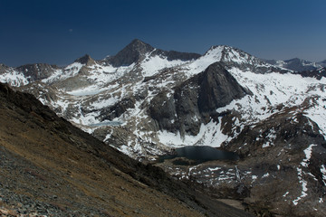 Obraz premium Snow covered peaks surround a hanging lake in the high sierra in California. A hiking trail descends the foreground mountainside