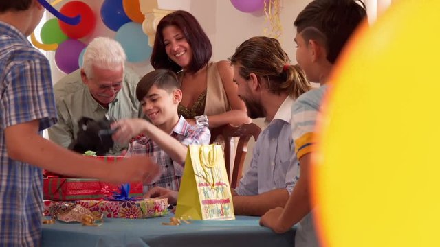 Group of happy children celebrating birthday at home, kids having fun at party. Boy opens present and shows toy to his family and friends. Slow motion