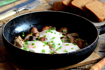 Fried eggs with mushrooms in a frying pan, brown bread slices, fork and knife on wooden table. Tasty and easy egg breakfast concept. Rustic style. Closeup
