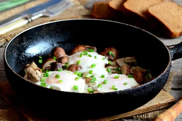Photo sur Aluminium Oeufs sur le plat Fried eggs with mushrooms in a frying pan, brown bread slices, fork and knife on wooden table. Tasty and easy egg breakfast concept. Rustic style. Closeup