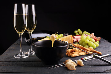 Gourmet Swiss fondue dinner on a winter evening with assorted cheeses on a board alongside a heated pot of cheese fondue with two forks dipping bread and white wine behind in a tavern or restaurant