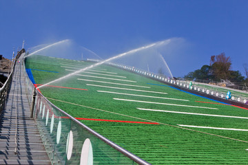 Fototapeta premium Green plastic coating of a ski jump hill is under summer maintenance with flushing water. Watering system at work