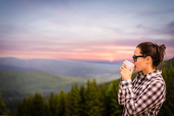 Young woman is enjoying cup of coffee outside with beautiful view of mountains and woods during a sunset/sunrise. Dark hair girl drinking cup of tea on a terace.