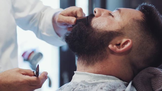 Senior handsome Italian barber is making a beard haircut to his hipster brutal bearded young customer. Italian barber traditions. Old fashioned male saloon. Inside shooting.