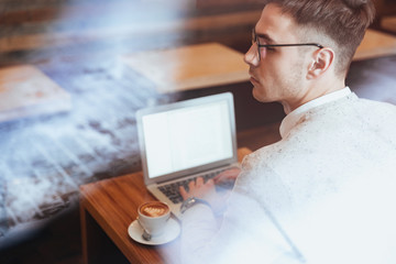 Young man wearing eyeglasses sitting in cafe and using laptop.