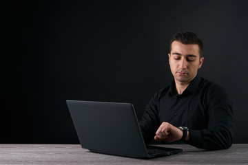 Man looking at clock and and working on a laptop. Dark background with copy space