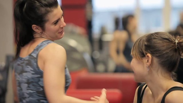 Two women relaxing and talking after workout in gym. Two girls talk in the gym. Girls laugh between exercise machines in the gym
