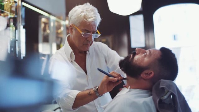 Skillful senior Italian barber is shaving his client with razor then checking the result. Popular Italian barberiano. Italian atmosphere, traditions. Typical barberiano design on the background.