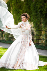 Bride in lace dress holds her veil on wind posing on green lawn