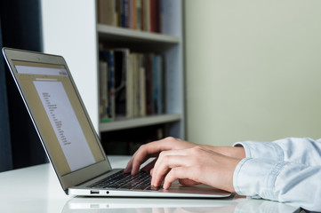 Working with laptop PC at home. Female hands type a list on computer at white minimalistic desk with book shelves in background