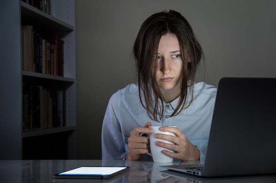 Tired Sleepy Female Person With Cup Of Coffee Working At Computer. Exhausted Female Person Holds Tea And Looks At Laptop Pc Screen At White Desk In Dark Room Late At Night