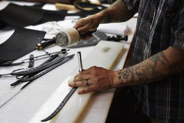 A man working at a bench in a leather workshop, using a mallet and chisel to mark leather. 
