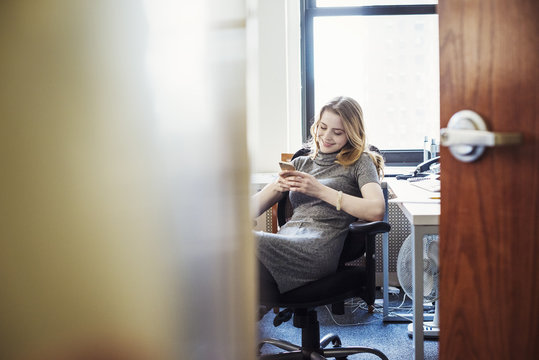 A woman in an office sitting checking her phone, seen through an open door. 