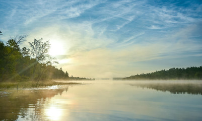 Fototapeta na wymiar Brilliant and bright mid-summer sunrise on a lake. Warm water and cooler air at daybreak creates misty fog patches. Still water along a calm, quiet Ontario lakeside. 