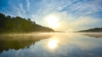 Fototapeta na wymiar Brilliant and bright mid-summer sunrise on a lake. Warm water and cooler air at daybreak creates misty fog patches. Still water along a calm, quiet Ontario lakeside. 