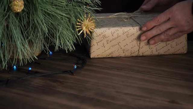The man puts box of gift under the Christmas tree. Fir stand on the wooden table and decorated with glowing garland and Christmas toys. Box with gift wrapped in brown paper and tied with twine.