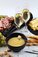Gourmet Swiss fondue dinner on a winter evening with assorted cheeses on a board alongside a heated pot of cheese fondue with two forks dipping bread and white wine behind in a tavern or restaurant