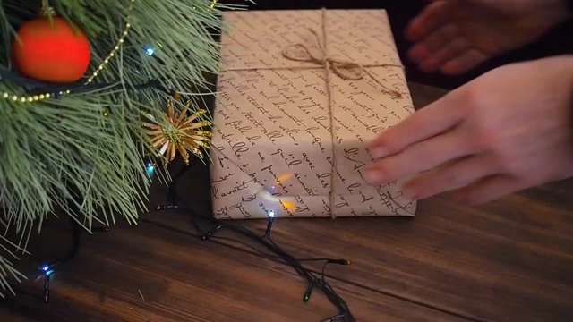 The man puts gift boxes under the Christmas tree. Fir stand on the wooden table, decorated glowing garland and Christmas toys. Boxes with gift wrapped in brown and colored paper and tied with twine.