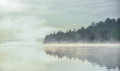 Fototapeta na wymiar Fog and mist rises all around, partially enshrouding a waterfront deciduous Eastern Ontario forest at a lakeside.