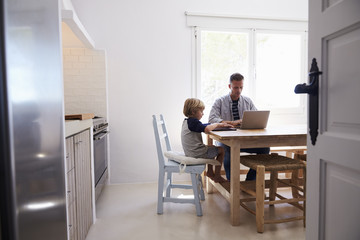 Dad and son using computers at kitchen table, from doorway