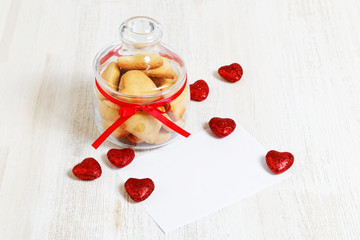 Fototapeta na wymiar Homemade heart shaped cookies in a glass jar, small red hearts and blank white mockup for greeting for Valentines Day on white wooden background