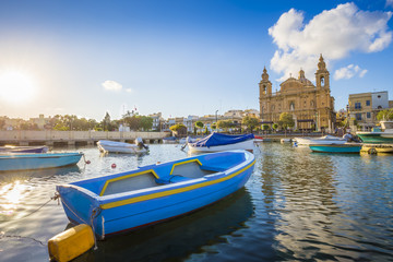 Fototapeta premium Msida, Malta - Blue traditional fishing boat with the famous Msida Parish Church at background on a summer day with blue sky and clouds