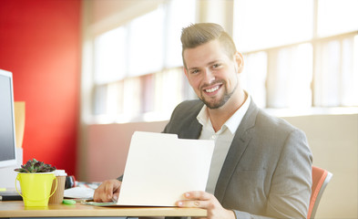 Confident male designer working and reviewing documents on a folder in red creative office space
