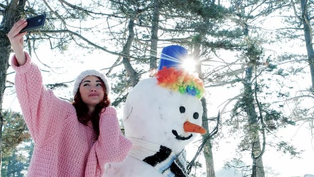winter selfie, cute girl making photo with snowman, a mobile phone in the hand of young woman making fun selfie photo in winter forest backlit, snow sculpture, fun entertainment