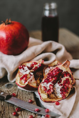 Big fresh juicy pomegranate and a half of one lie on the old crumpled kitchen towel, on the rough wooden table, old knife, glass bottle with tasty pomegranate juice and the dark gray background