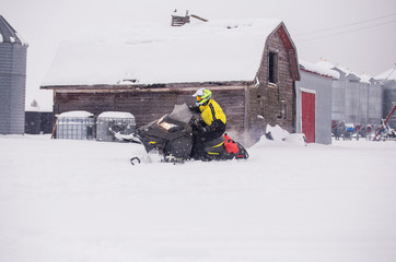 A man in motion tipping his snowmobile on the side for fun with an old barn in the background in a farm yard landscape