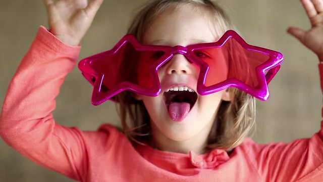 Funny little girl in big glasses in the shape of stars looks at the camera smiles and shows his tongue and his hands. Funny little girl in pink glasses looks at the camera and grimaces