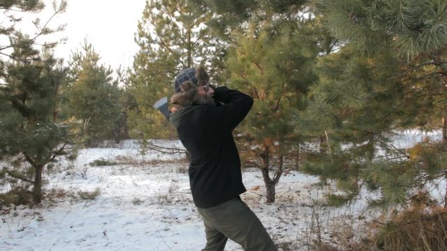 Strange man in a hipster sunglasses with beard throws an ax to a tree. Woman in a hat with ear flaps walking in the forest.