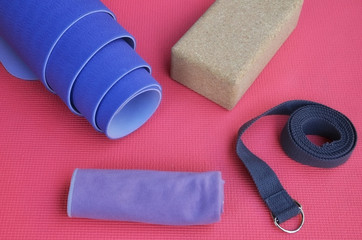 Yoga Accessories: rolled lilac exercise mat, cork block, grey strap and towel on bright pink yoga mat background.