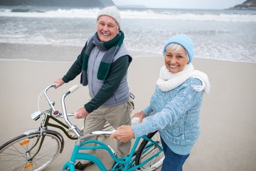 Portrait of senior couple standing with bicycles on the beach  - obrazy, fototapety, plakaty