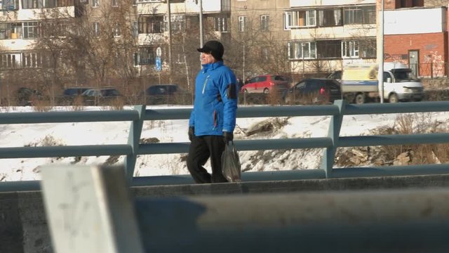 Urban passer. Winter.

A man of mature age in a blue warm jacket with the package goes against the backdrop of passing vehicles.