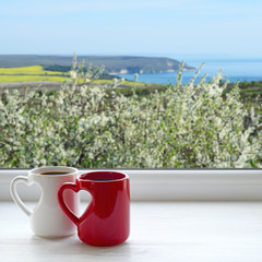 Two coffee cups on a windowsill. Outside, flowering trees, fields, mountains and the sea