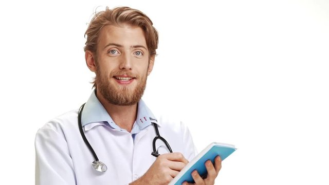 Glad Caucasian male doctor with light beard wearing medical overall smiling making notes in blue notebook on white background in slowmotion