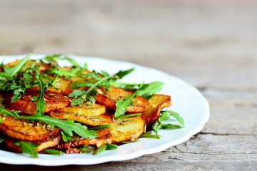 Crispy roasted potatoes with arugula on a white plate. Wooden background. Easy vegetable side dish made with potatoes and arugula. Warm salad. Vegetarian food. Closeup