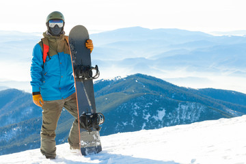 Young snowboarder standing next to snowboard thrusted into snow with beautiful scenery on background - obrazy, fototapety, plakaty