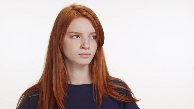 Outraged red-haired Caucasian teenage girl standing on white background looking at camera and away