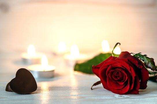 Red rose lying on white a wooden surface. In the background are lit small candles. Next to the flower is two chocolates in the shape of hearts.