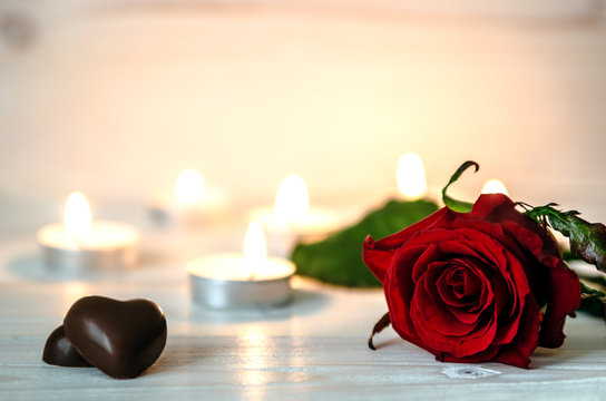 Red rose lying on white a wooden surface. In the background are lit small candles. Next to the flower are chocolates in the shape of hearts.