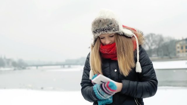 Girl taking smartphone from the jackt's pocket while standing next to the frozen river
