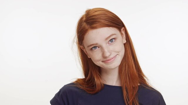 Smiling charming Caucasian teenage ginger girl in dark blue t-shirt standing on white background blinking fast